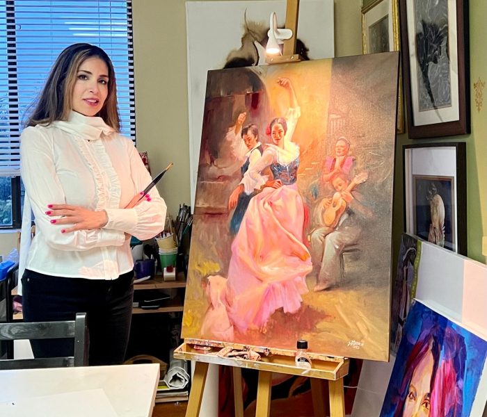 A woman in a white blouse and black pants stands beside an easel holding a vibrant oil painting of flamenco dancers performing with musicians. The studio is filled with art supplies and framed portraits.